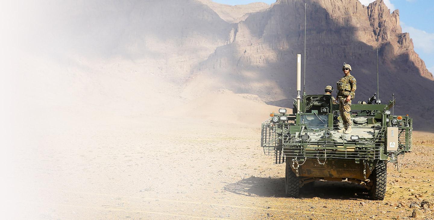 Military man wearing combat gear standing on the front of a green army vehicle against a rocky mountain backdrop